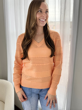Woman wearing an orange sweater and blue jeans standing in a room with white curtains.