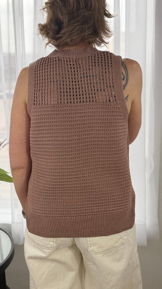 Person wearing a brown knitted vest standing in front of white curtains