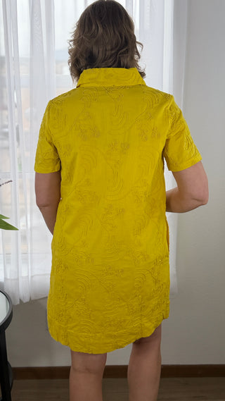 Person wearing a yellow dress with a subtle pattern in a room with white curtains.