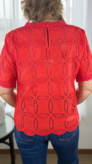 Person wearing a red lace top with blue jeans indoors.
