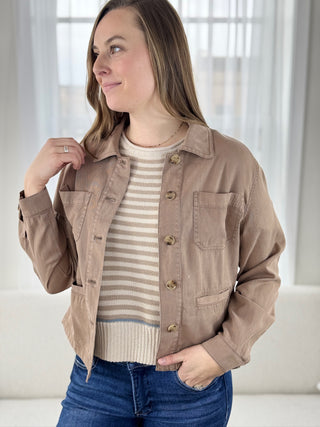 Woman wearing a brown jacket over a striped shirt with blue jeans indoors.