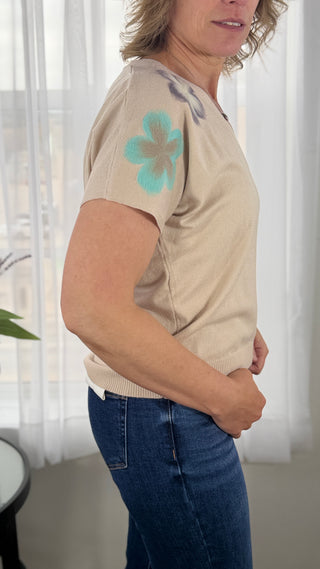 Woman wearing a beige t-shirt with a blue flower design in a room with white curtains.