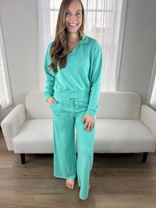 Woman wearing a teal tracksuit standing in a room with a white couch and light-colored walls.