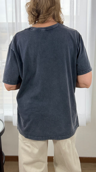 Person wearing a dark blue t-shirt and light-colored pants standing in front of a window with white curtains.