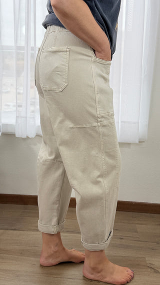 Person wearing beige pants standing indoors with white curtains in the background