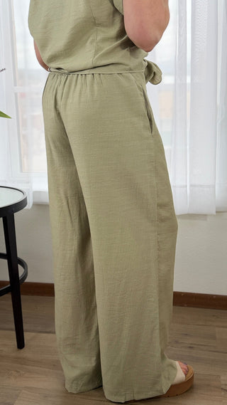Person wearing a green jumpsuit standing indoors with white curtains in the background