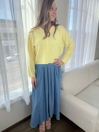 Woman wearing a yellow blouse and blue skirt standing in a room with white curtains.