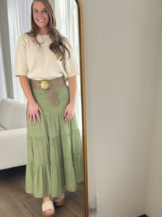 Woman wearing a green tiered skirt and beige top in a room with a couch and mirror.