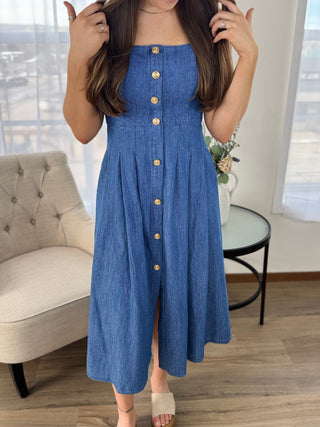 Woman wearing a blue denim dress with gold buttons in a room with a couch and table.