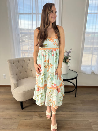 Woman wearing a floral dress standing in a room with a chair and table.