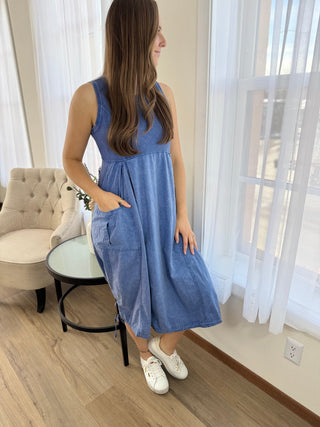 Woman wearing a blue dress standing in a room with light-colored walls and wooden flooring.