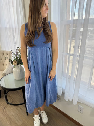 Woman wearing a blue dress standing in a room with white curtains and a table.