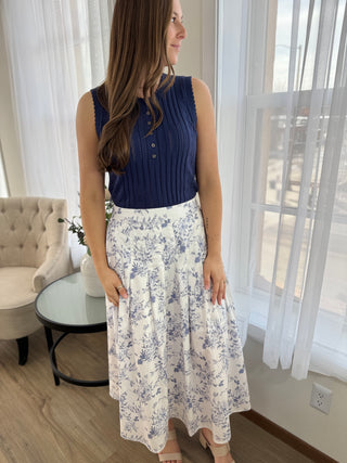 Woman wearing a blue top and floral skirt in a room with white curtains and a chair.