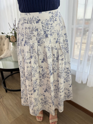 Person wearing a floral skirt in a room with white curtains and a table.