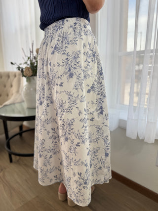 Person wearing a floral skirt in a room with light curtains and a table.
