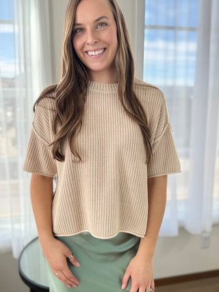 Woman wearing a beige knitted top in a bright room with large windows.