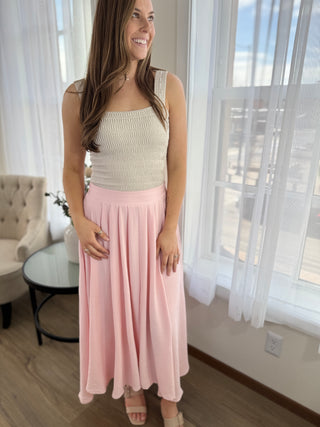 Woman wearing a beige top and pink skirt in a room with large windows.