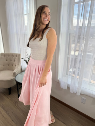 Woman wearing a sleeveless top and pink skirt standing in a room with large windows.