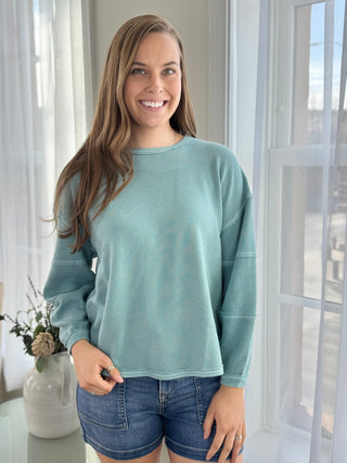 Woman wearing a light blue sweater and denim shorts standing in a bright room with sheer curtains.