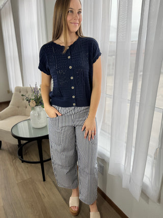Woman wearing a navy blue top and striped pants standing in a room with light curtains.