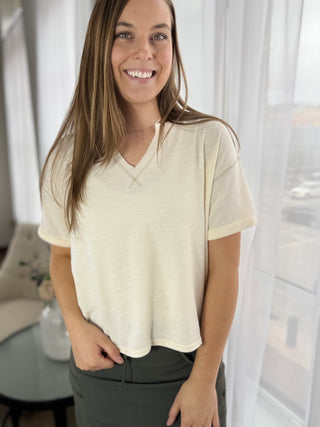 Woman wearing a beige shirt and gray pants indoors with white curtains in the background