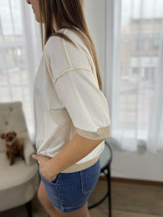 Woman wearing a white blouse with beige accents and blue shorts in a room with large windows.