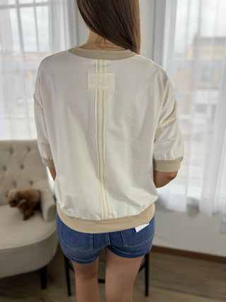 Person wearing a beige top with a visible brand label in a room with a couch and teddy bear.