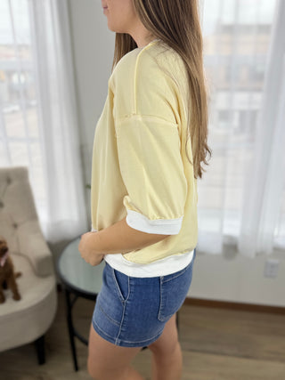 Person wearing a yellow top and denim shorts in a room with white curtains.