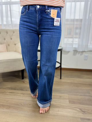 Person wearing blue jeans with a visible brand tag in a room with a couch and window blinds.
