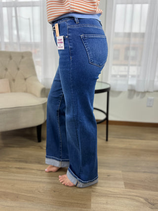 Person wearing blue jeans standing in a room with a couch and window blinds.
