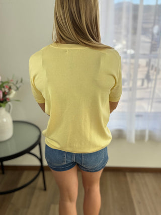 Person wearing a yellow top and denim shorts standing in a room with a window in the background.