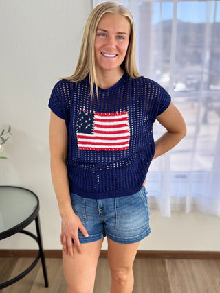 Woman wearing a navy blue knit top with an American flag design in a room with a window in the background.