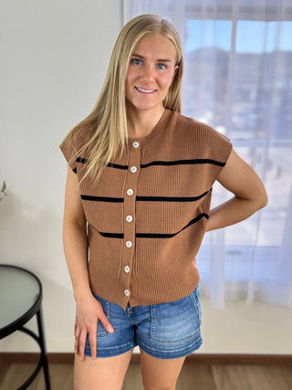 Woman wearing a brown knitted vest with black stripes and denim shorts indoors.