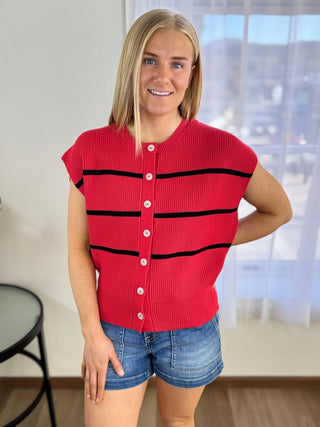 Woman wearing a red sleeveless sweater with black stripes and denim shorts indoors.