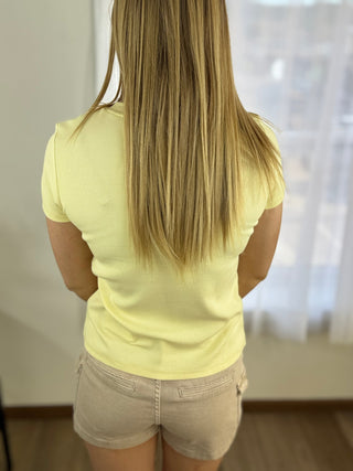 Person wearing a yellow t-shirt and beige shorts standing in a room with a window in the background.