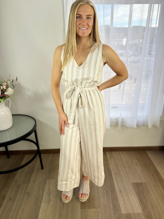 Woman wearing a white striped jumpsuit standing in a room with a table and window in the background.