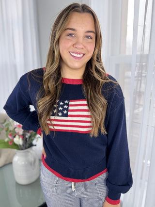 Woman wearing a navy sweater with an American flag design, standing indoors.