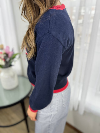Person wearing a navy sweater with red trim, standing in a room with white curtains and a table with flowers.