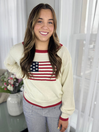 Woman wearing a cream sweater with an American flag design, standing indoors.