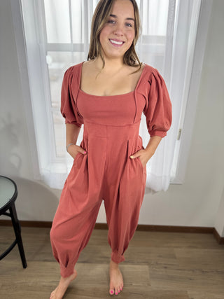 Woman wearing a pink jumpsuit standing in a room with white curtains.