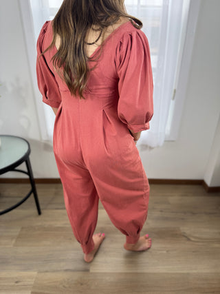 Person wearing a pink jumpsuit standing in a room with white curtains and a wooden floor.