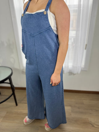 Person wearing blue denim overalls standing indoors with white curtains in the background