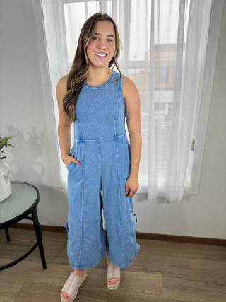 Woman wearing a blue jumpsuit standing in a room with white curtains.