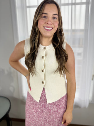Woman wearing a cream sleeveless top with buttons and a pink skirt indoors.
