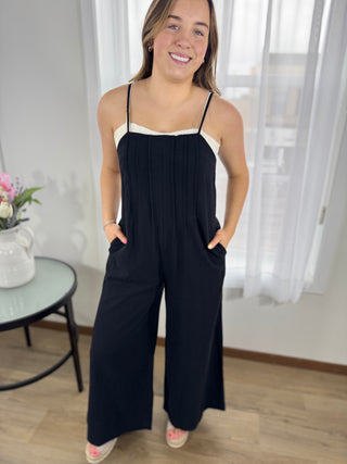 Woman wearing a black and white jumpsuit standing in a room with light-colored walls and wooden floor.