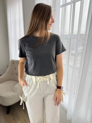 Woman wearing a dark gray t-shirt and light-colored pants standing in a room with white curtains.