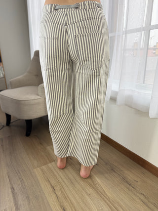 Person wearing striped pants standing in a room with a chair and window in the background.