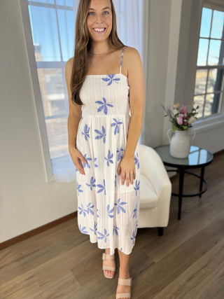 Woman wearing a white dress with blue floral patterns in a room with large windows.