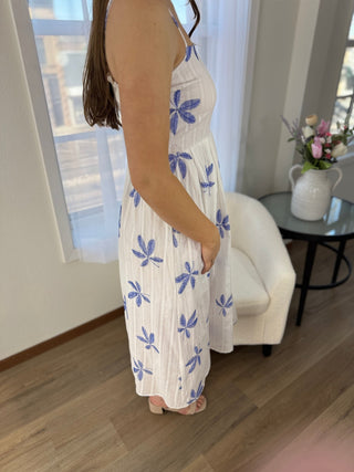 Woman wearing a white dress with blue floral patterns in a room with a white chair and a small table with a vase.
