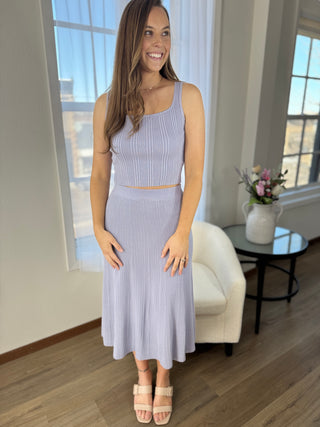 Woman wearing a light purple ribbed dress standing in a room with large windows.